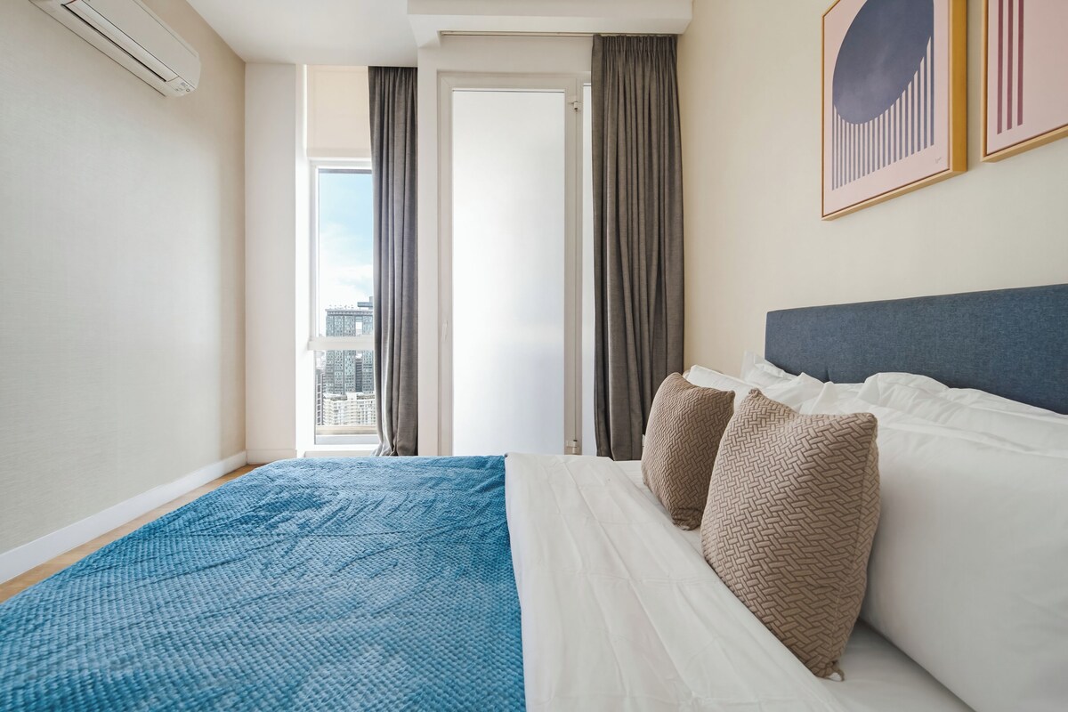 Ein Hotelzimmer mit Bett, einem fenster mit Vorhängen, Blick auf die Stadt und einem gerahmten Bild an der Wand.