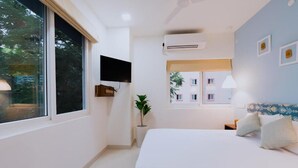 Un dormitorio con cama, televisor montado en la pared, equipo de aire acondicionado, ventana y una planta.