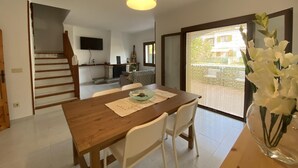 A dining area with a wooden table, white chairs, a hanging light fixture, and a view through a glass door. - HOUSE WITH COMUNITY POOL (l'Escala)