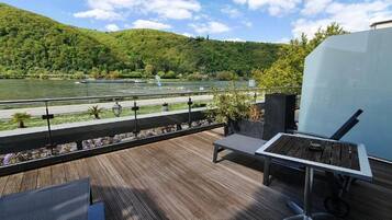 Una terraza con vista a un río y colinas, mobiliario exterior y un gran muro blanco.