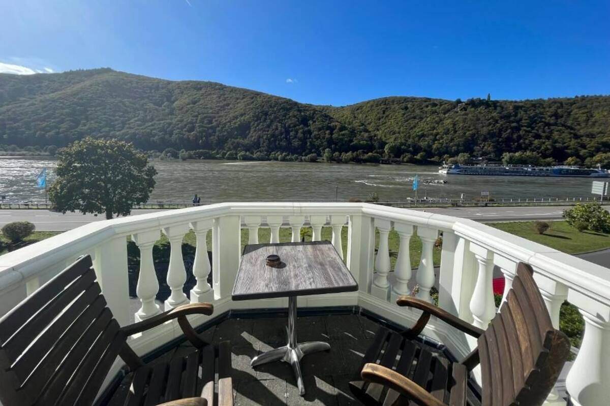 A balcony with a table and chairs overlooking a lake with boats and hills in the background.