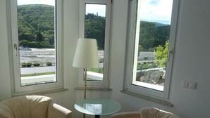 A room with two beige armchairs, a small glass table with a white lamp, and large windows offering a view of a river and greenery. - Hotel Schön (Ruedesheim am Rhein)