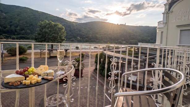 A balcony with a cheese board, a glass of wine, and a view of a lake and hills. - Hotel Schön (Ruedesheim am Rhein)