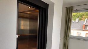 A room with a sliding door leading to a metallic elevator, a window with curtains, and a view of buildings outside. - Hotel Schön (Ruedesheim am Rhein)