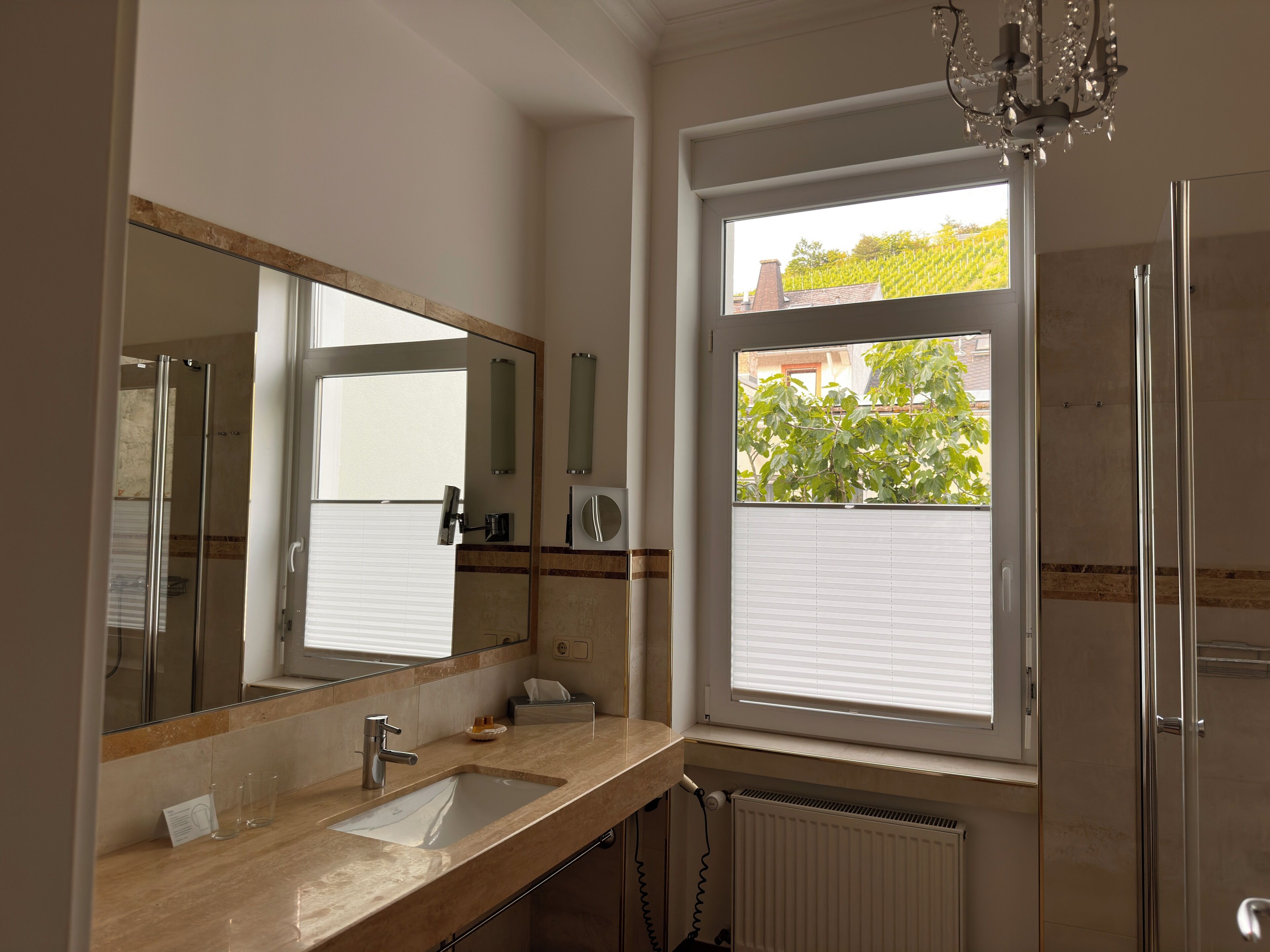 Une salle de bain comprenant un grand miroir, un lavabo, un distributeur de serviettes, une fenêtre avec des stores et un espace douche.