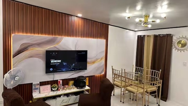 A modern living room with a flat-screen TV mounted on a wall, a dining area with a glass table and chairs, and a ceiling fan.