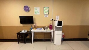 Quarto com televisão de tela plana, uma mesinha com cadeira, geladeira e relógios fixados na parede.