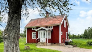 A red wooden house with a white door and windows, a gravel driveway, and a large tree in the foreground. - 4 Person Holiday Home in Aneby (Aneby)