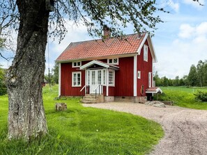 Ein rotes Holzhaus mit weißer Tür und Fenstern, einer Schotterauffahrt und einem großen Baum im Vordergrund.