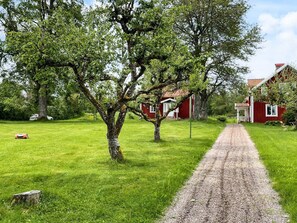 Ein Kiesweg führt zu einem roten Gebäude mit einer weißen Tür, umgeben von üppigem Grün und Bäumen.