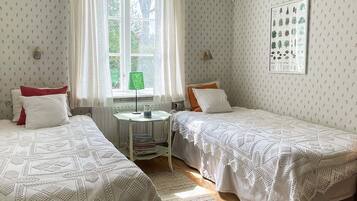 A room with two beds, a window with curtains, a small table with a lamp, and a wall-mounted plant.
