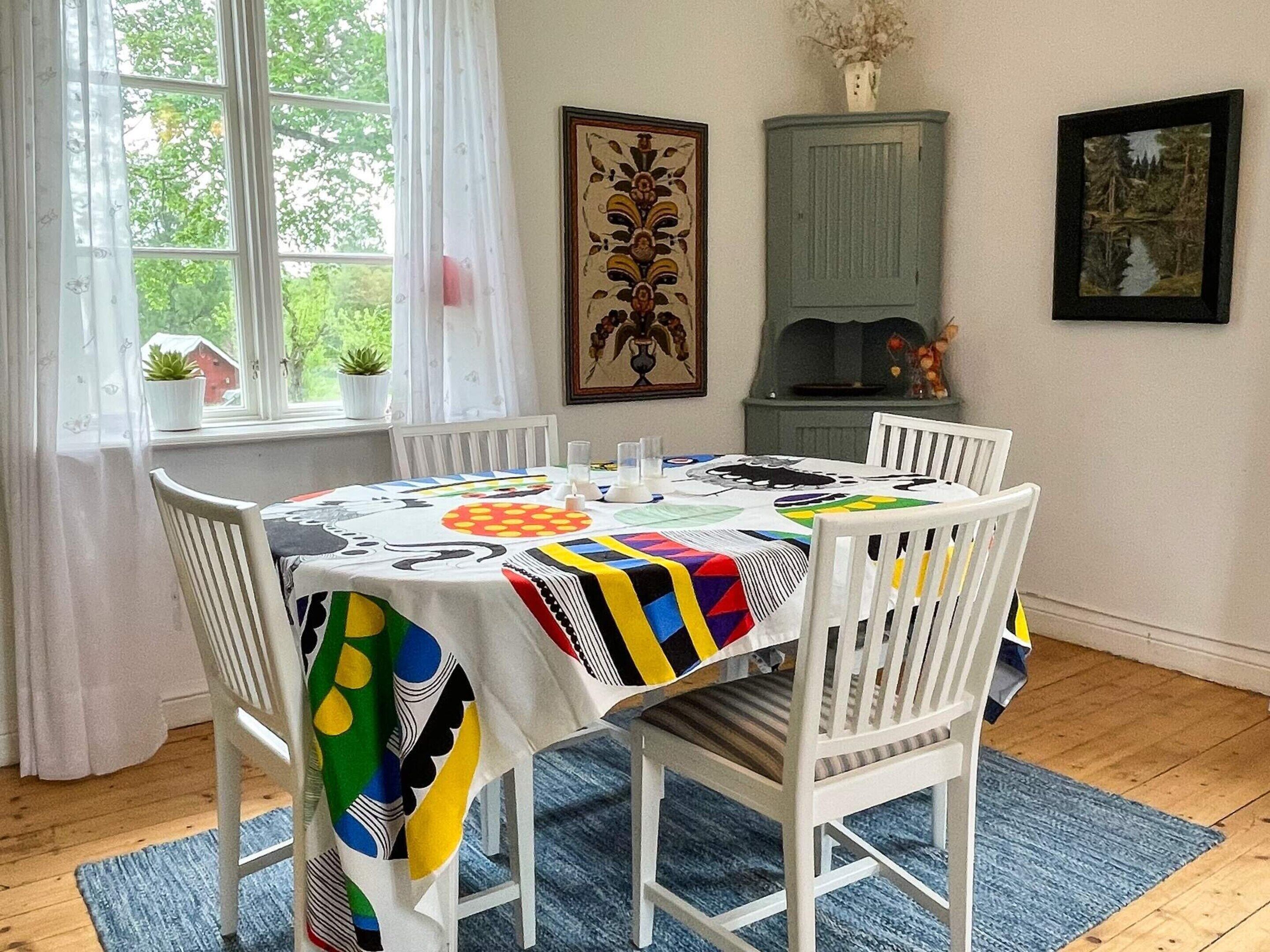 A dining area with a table set for four, white chairs, a colorful tablecloth, and a window with curtains.