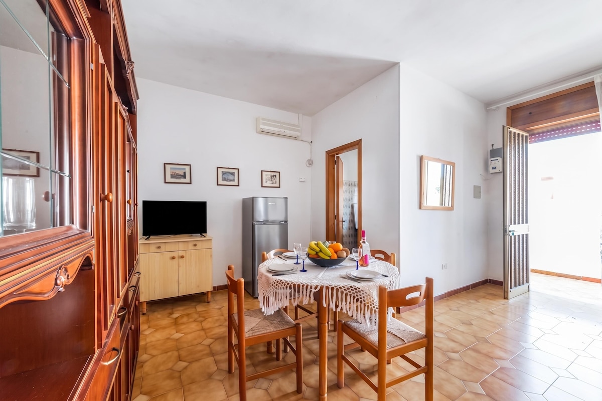 A dining area with a table set for two, a refrigerator, and a television.