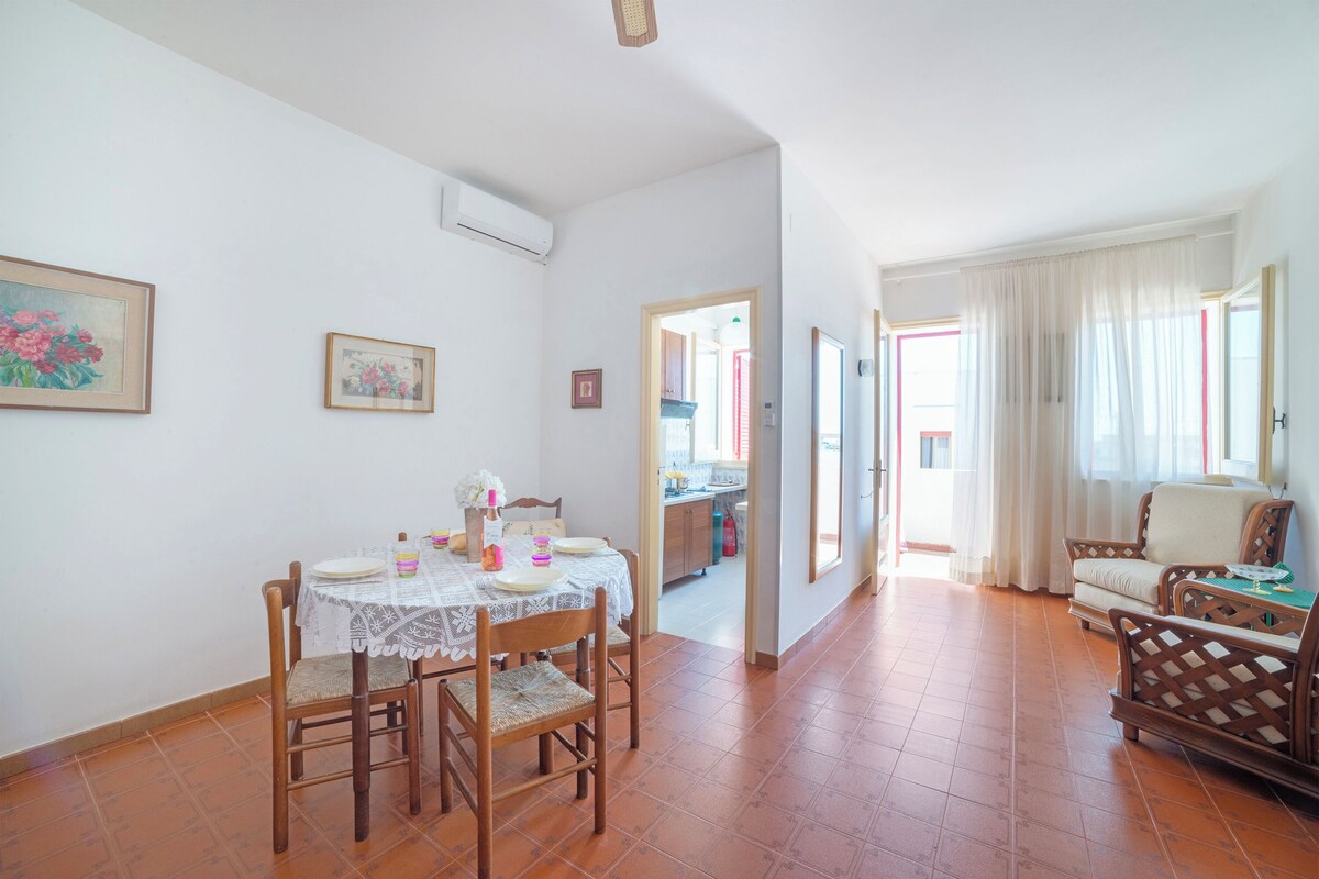 A dining area with a table set for four, a kitchen in the background, and a living area with a sofa and armchairs.