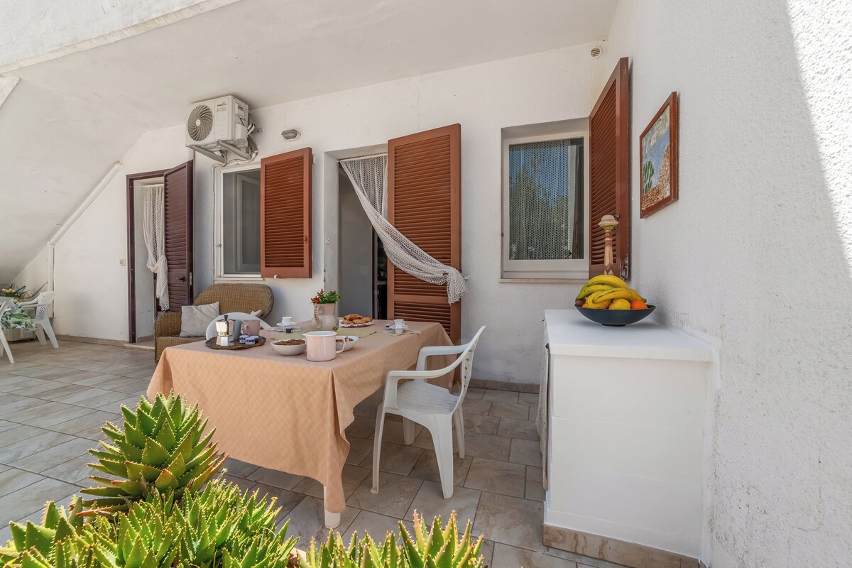 A dining area with a table set for two, a white chair, a small white cabinet with a bowl of fruit, and a window with brown shutters.