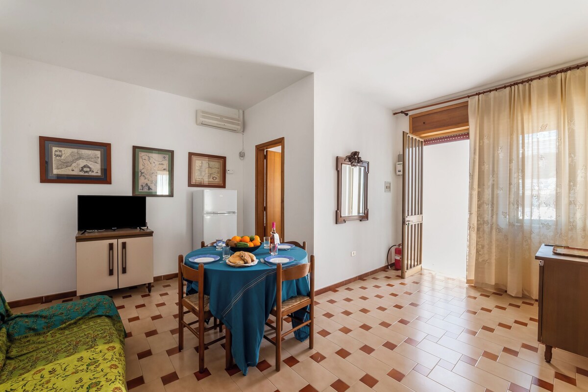 A dining area with a table set for two, a television on a cabinet, and a door leading to another room.