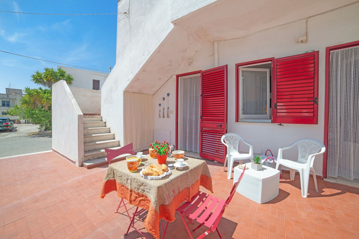 A terrace with a table set for dining, red shutters, and a staircase leading to an upper level.
