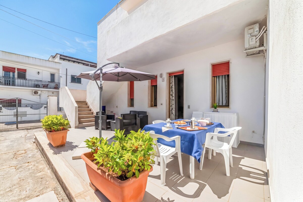 An outdoor patio with a table set for dining, surrounded by potted plants and white chairs.