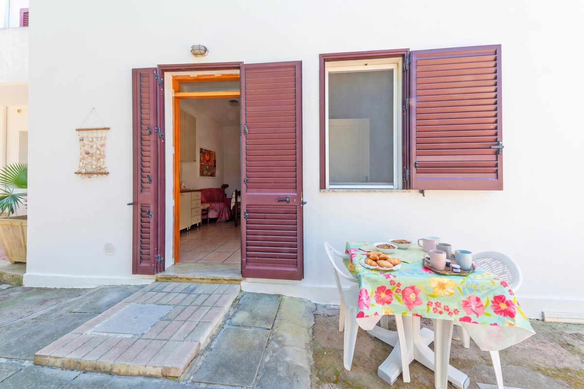 A white building with a door open leading to an interior room, a window with shutters, and a table with a floral tablecloth outside.