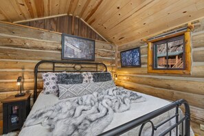 A log cabin bedroom with a bed, framed pictures, a nightstand, and a window with a view.