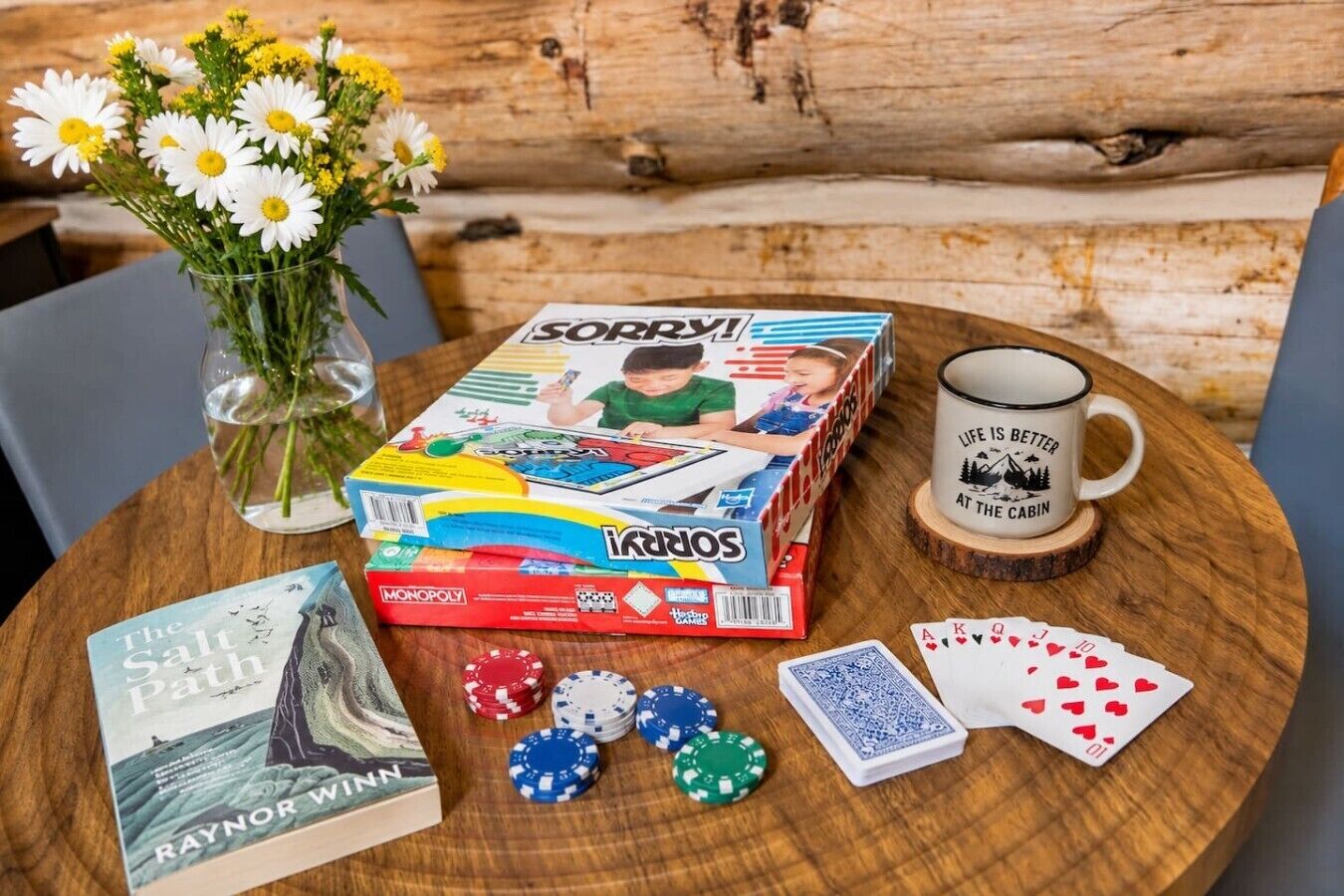 Ett Monopolyspel upplagt på ett träbord, tillsammans med en bok, pokermarker, spelkort, en mugg och en vas med blommor.