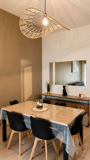 Ein Essbereich mit Holztisch, schwarzen Stühlen, einer gewebten Pendelleuchte und einer Küche im Hintergrund.