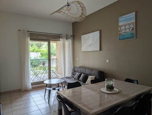 Ein Zimmer mit Esstisch, einer Couch, einem kleinen Tisch mit Stühlen, einem Deckenventilator, einem Fenster mit Vorhängen und einem an der Wand hängenden Bild.