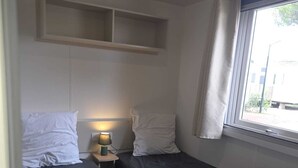 A compact room with two beds, a small table with a lamp, a window with curtains, and a shelf above the beds. - Air-conditioned mobile home for 6–8 people—direct beach access at a 4-star comfort campground (Canet-en-Roussillon)