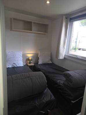 A compact room with two beds, a small table with a lamp, a window with curtains, and a shelf above the beds. - Air-conditioned mobile home for 6–8 people—direct beach access at a 4-star comfort campground (Canet-en-Roussillon)