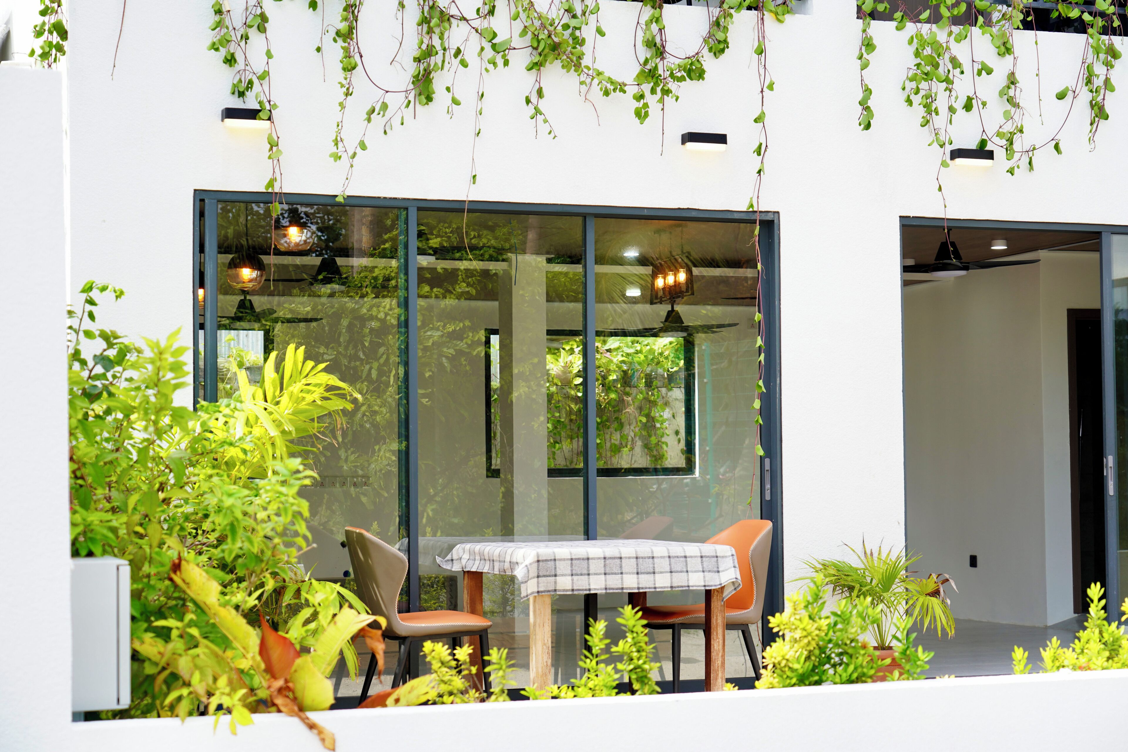 A modern outdoor dining area with a table and chairs, surrounded by lush greenery and large windows.