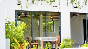 A modern outdoor dining area with a table and chairs, surrounded by lush greenery and large windows.