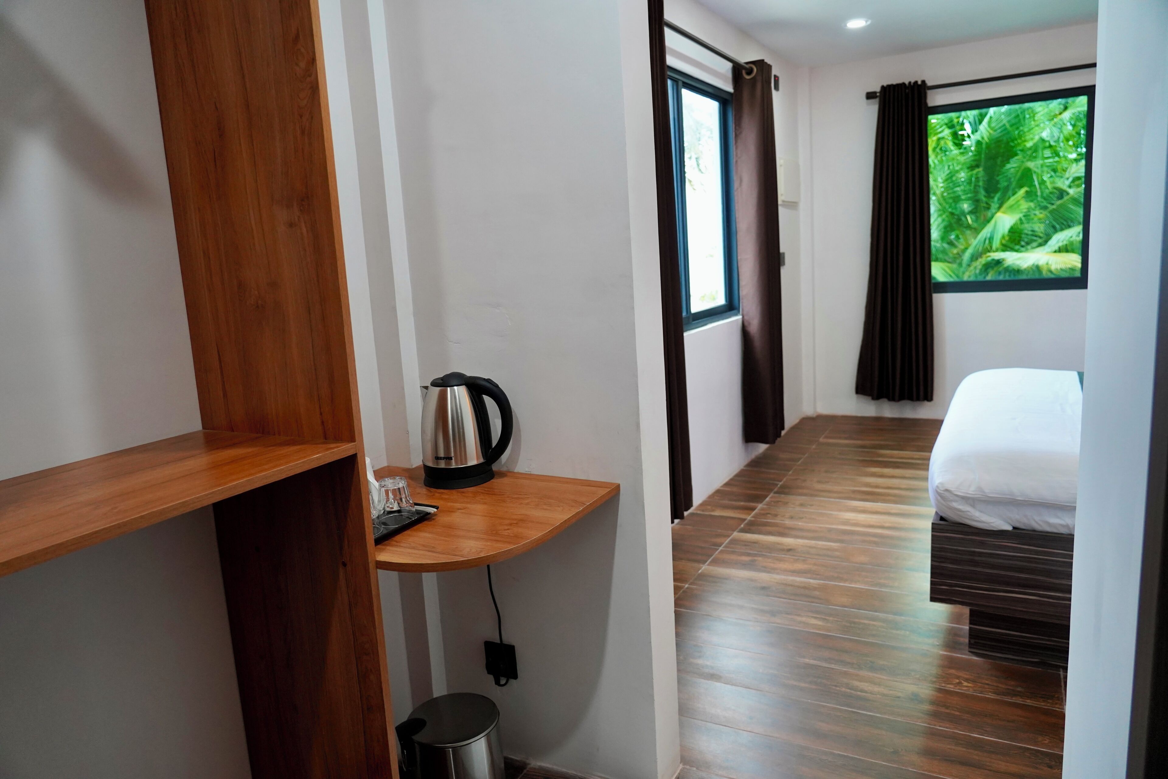 A hotel room with a bed, a small table with a kettle, a trash bin, and a window with curtains.