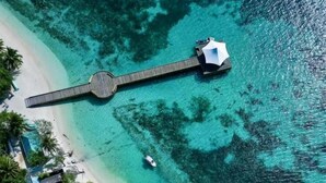 Vista aérea de una playa con un muelle de madera que conduce a una estructura blanca, probablemente un hotel o resort.