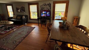 A living room with a black leather sofa, a wooden dining table with chairs, a TV on a stand, and a large window with blinds.