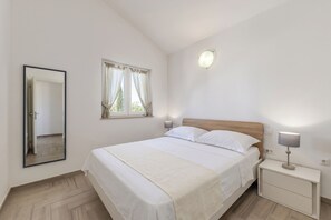 A bedroom with a bed, a nightstand, a wall-mounted mirror, a window with curtains, and a ceiling light. - Wabi Sabi Apartment 7 (Krk)