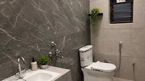A modern bathroom with a marble wall, a white sink, a toilet, a shower, and a small shelf with a plant.
