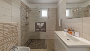 Et moderne badeværelse med bruser, håndvask og toilet. Et lille vindue er placeret over bruseren, og væggene er beklædt med fliser.