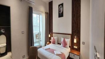 A hotel room with a bed, a chair, a desk, a window with curtains, and an air conditioning unit.
