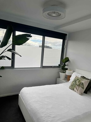 Un dormitorio con un ventanal grande, una cama, una mesita de noche con lámpara y una planta en maceta.