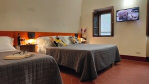 A hotel room with two beds, a TV mounted on the wall, a window, and a nightstand with a bottle and glasses. - A comfortable standard triple room just a stone's throw from the center of Volterra (Volterra)