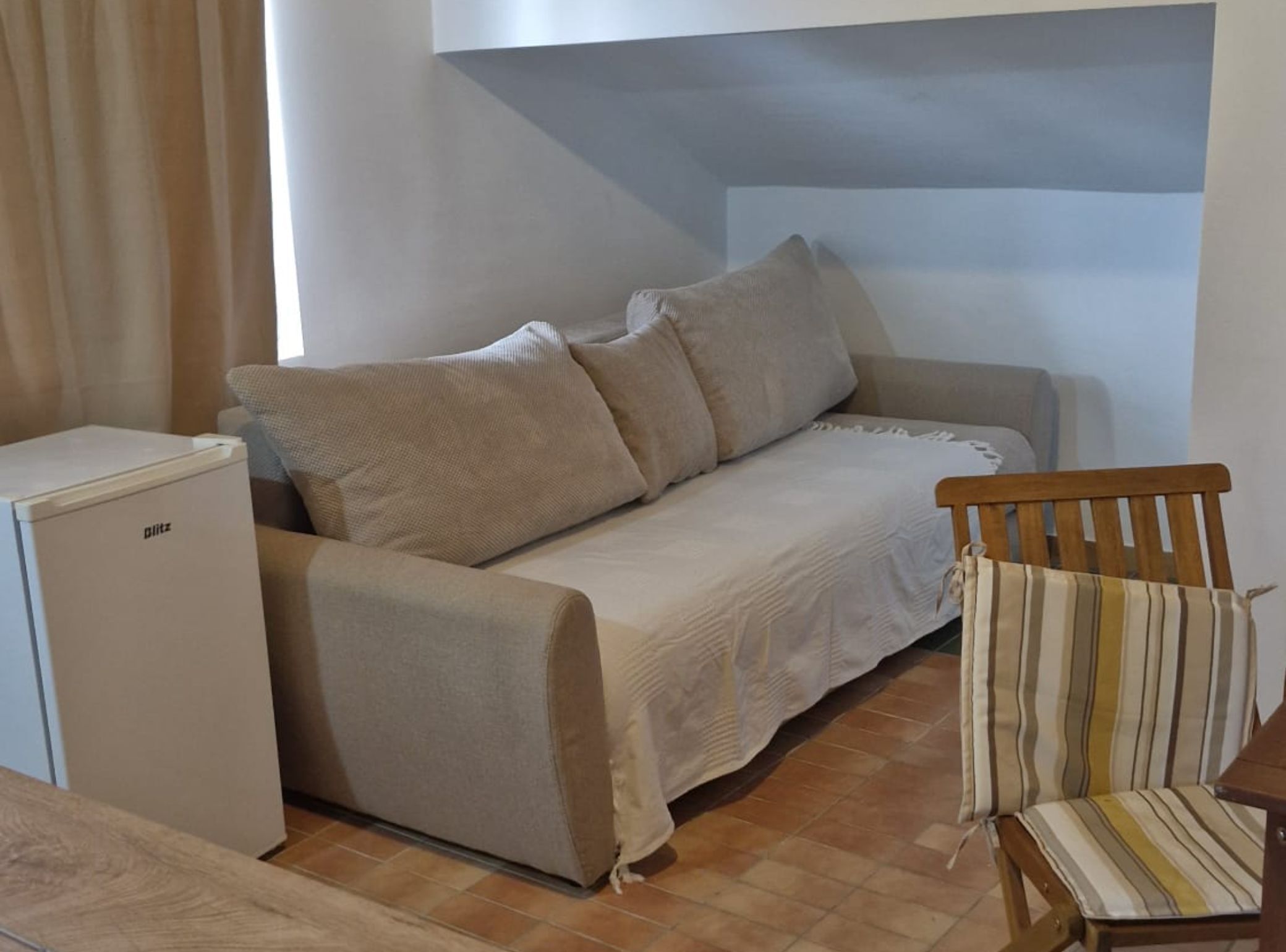 A small, cozy room with a sofa, a wooden chair, and a compact refrigerator.