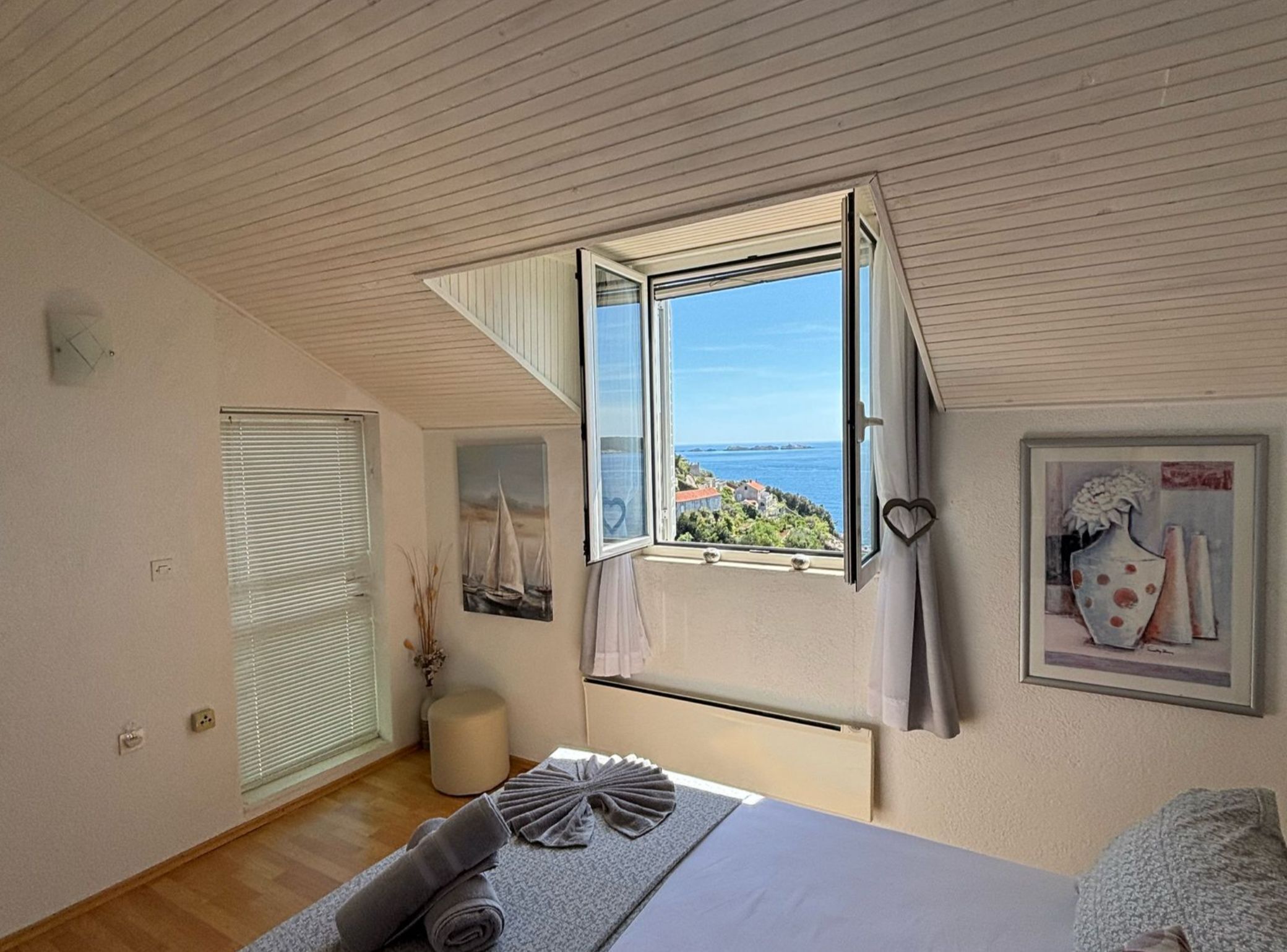 Un dormitorio con un ventanal grande, una cama, una mesita de noche, un taburete pequeño y un cuadro en la pared.