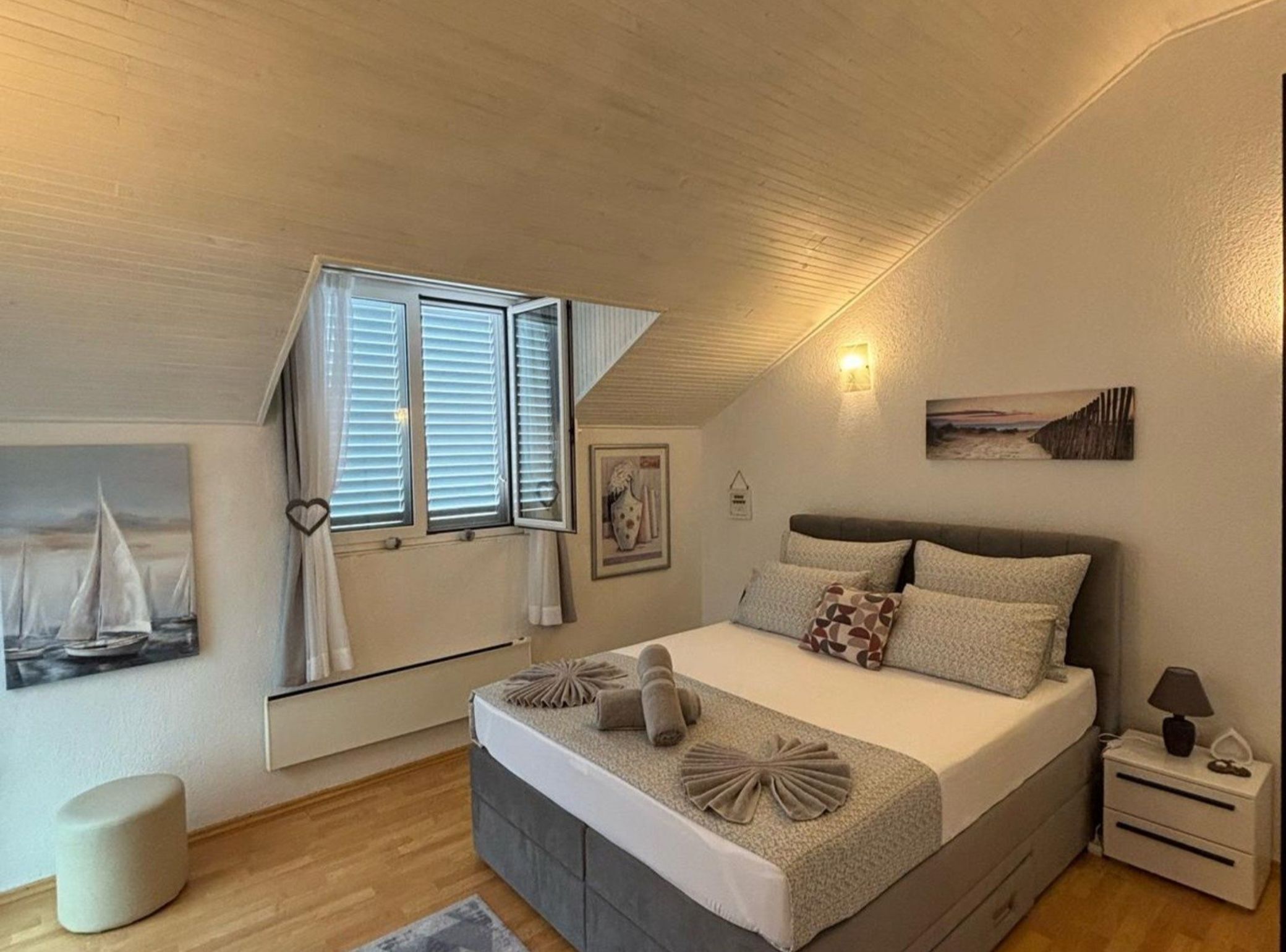 A bedroom with a bed, a nightstand, a lamp, a window with curtains, a wall-mounted picture, and a small stool.