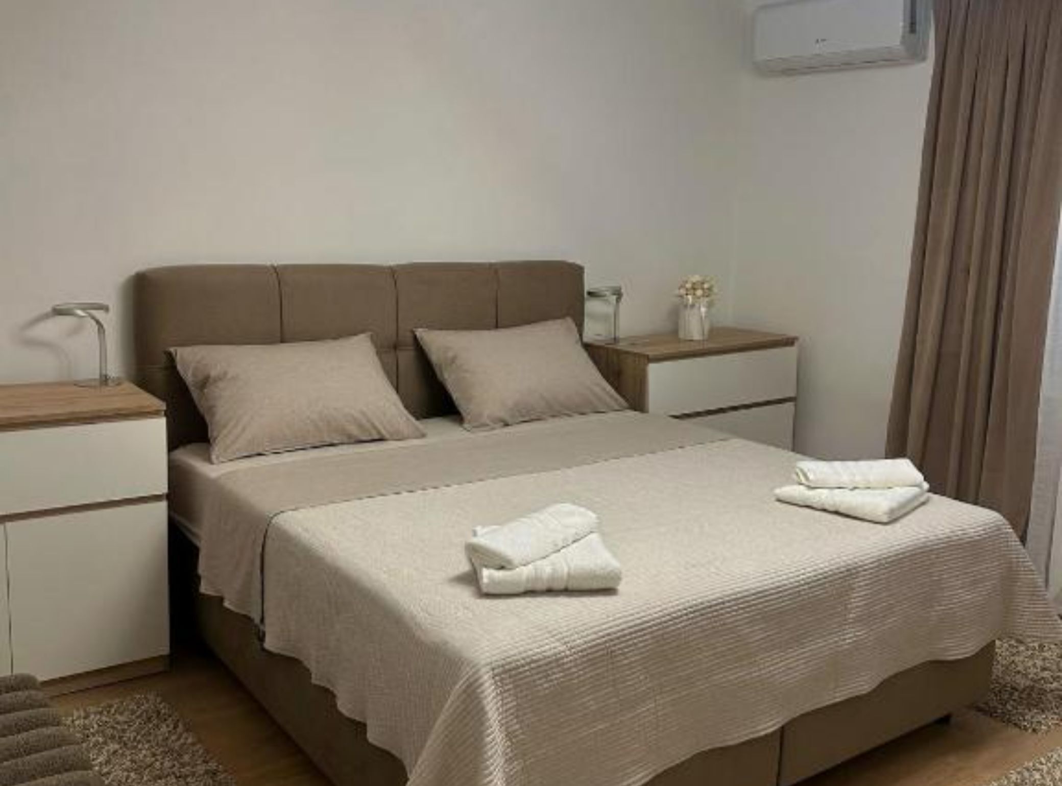 A bedroom with a bed, two bedside tables, a nightstand, a lamp, and a wall-mounted air conditioner.
