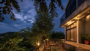Une terrasse extérieure avec une table et des chaises, entourée de plantes en pot et d’un arbre.