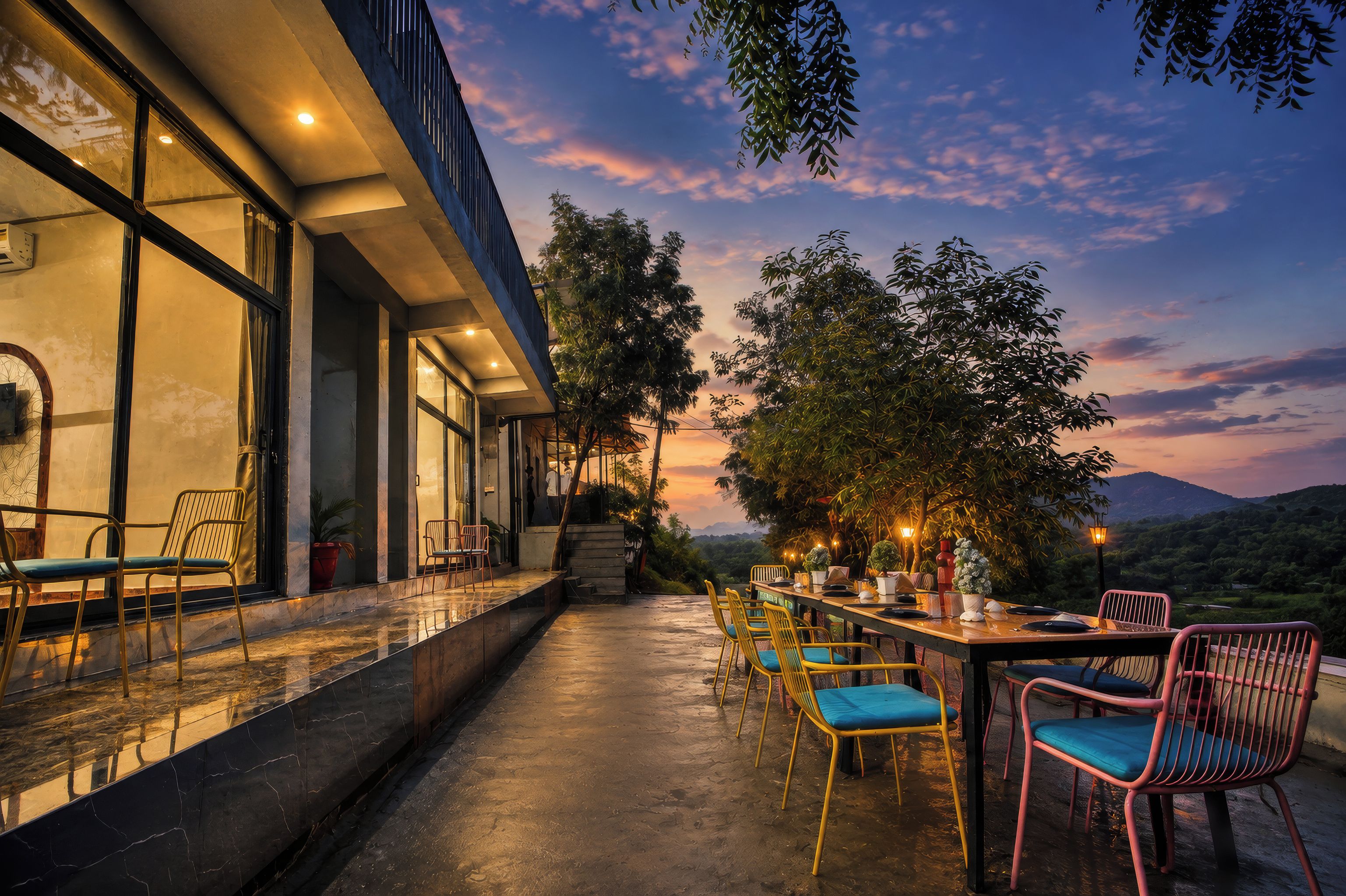An outdoor dining area with tables and chairs, a view of the landscape, and a building with large windows.