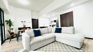 Ein modernes Wohnzimmer mit einer Eckcouch, einem Essbereich mit Tisch und Stühlen, einer Küchenzeile mit Schränken und Geräten sowie einem großen Fenster.