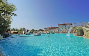 A large outdoor swimming pool with water slides and rock formations, surrounded by umbrellas and a building with a red roof. - Piani Di Clodia 2 p4p Clim (Lazise)