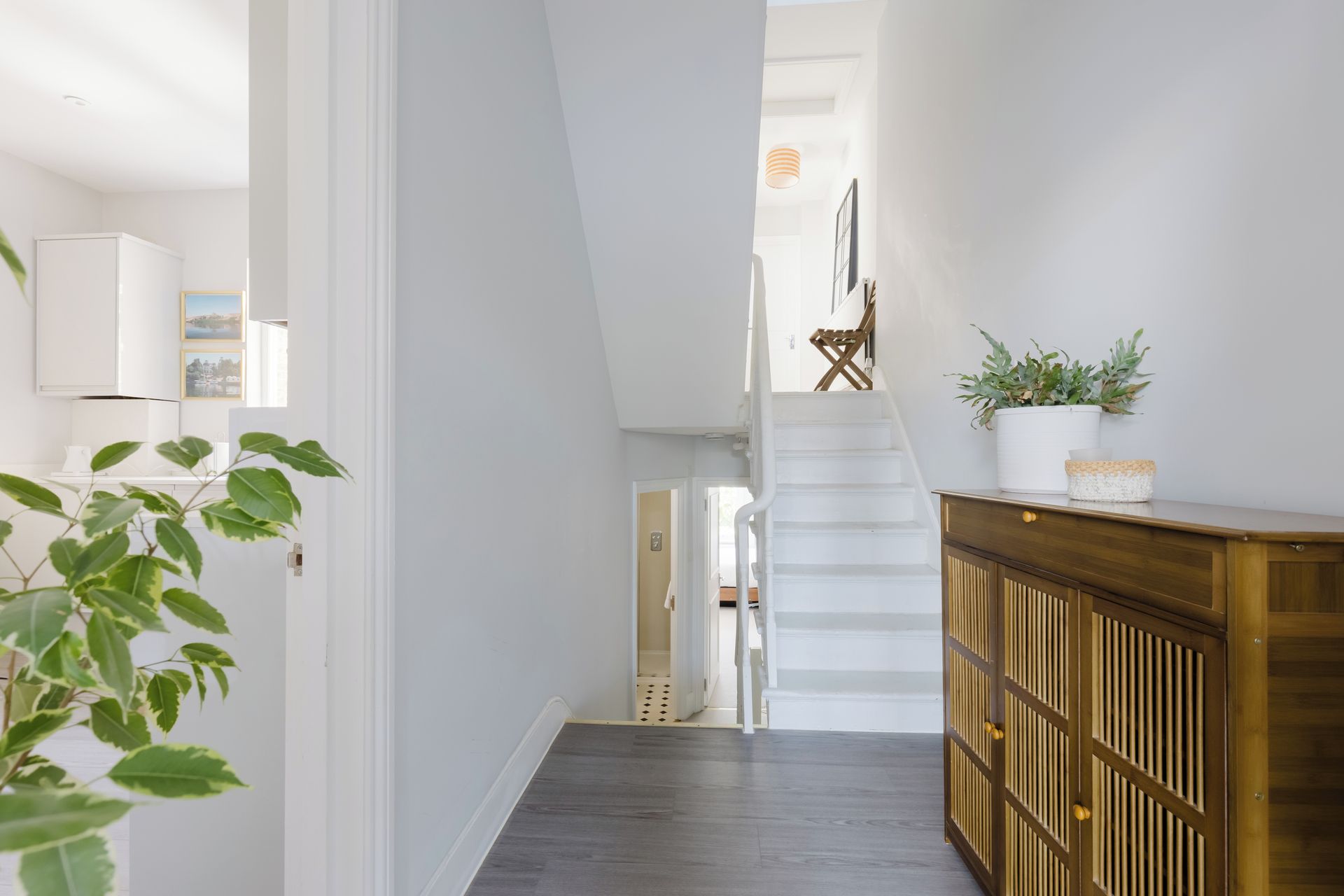 A modern interior with a staircase leading to an upper level, a wooden cabinet with a plant, and a white wall with a picture frame.