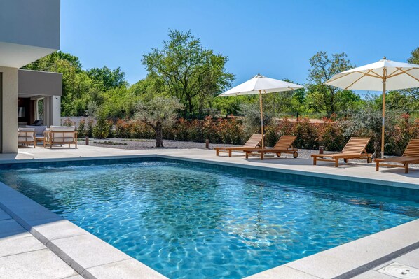 A rectangular swimming pool with clear blue water, surrounded by lounge chairs and umbrellas, and a modern building in the background. - Nareo Villa Collection With Private Pool (Sveta Nedelja)